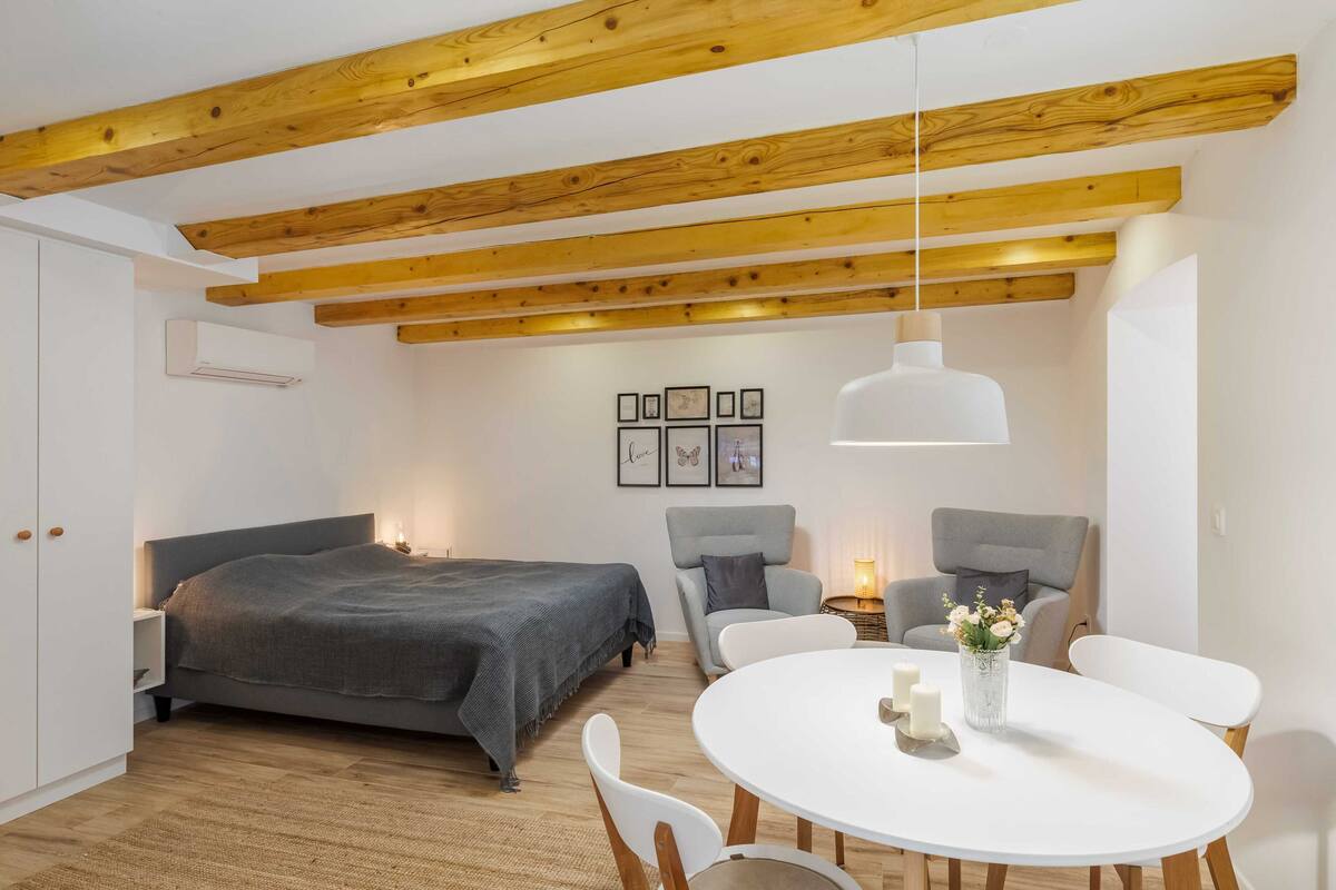 A bedroom with a bed, bedside tables, a round dining table with chairs, a hanging light fixture, and a wall-mounted air conditioner.