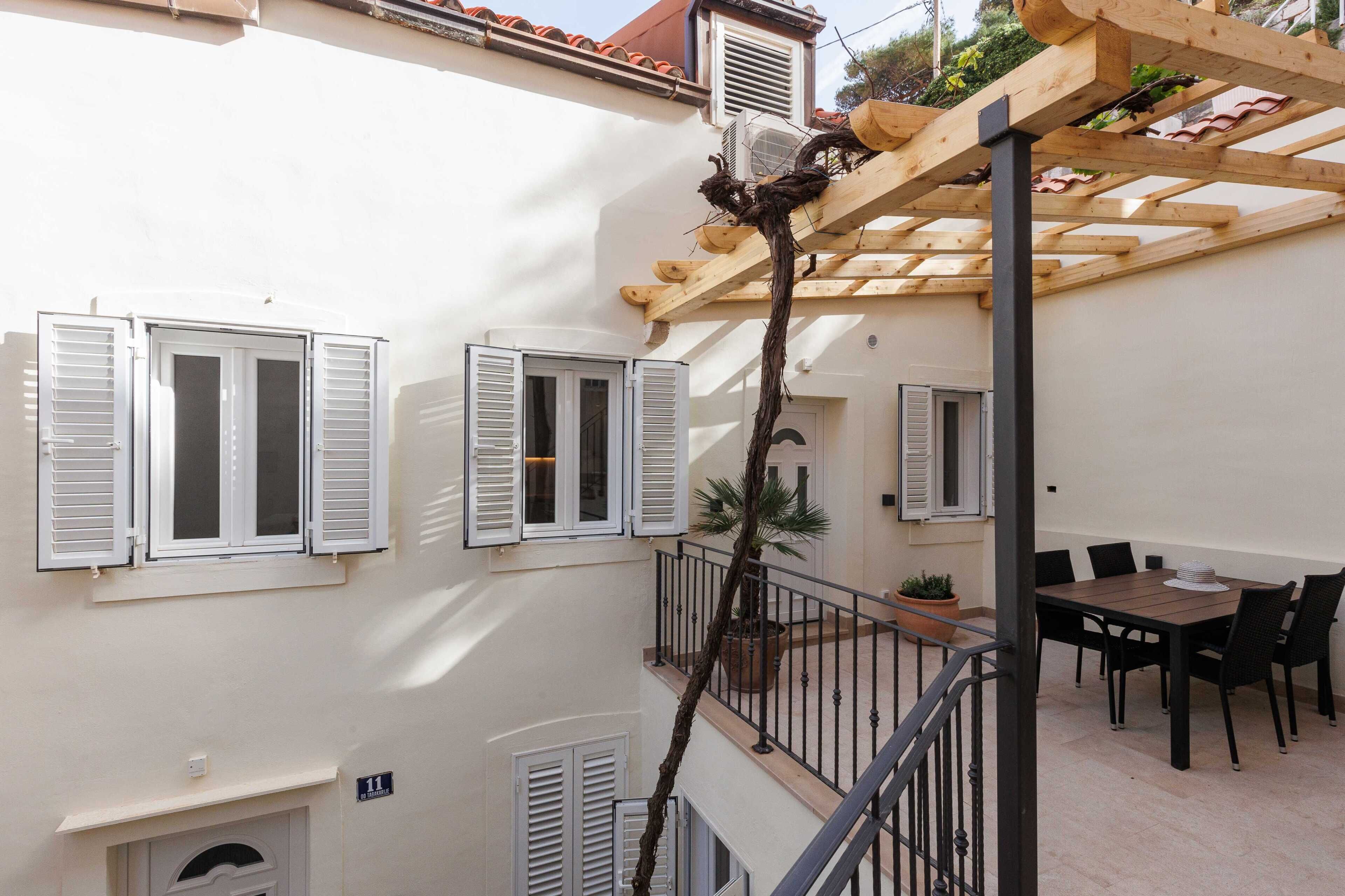 Un edificio de dos pisos con paredes blancas y contraventanas, una pérgola de madera y un balcón con mesa y sillas.