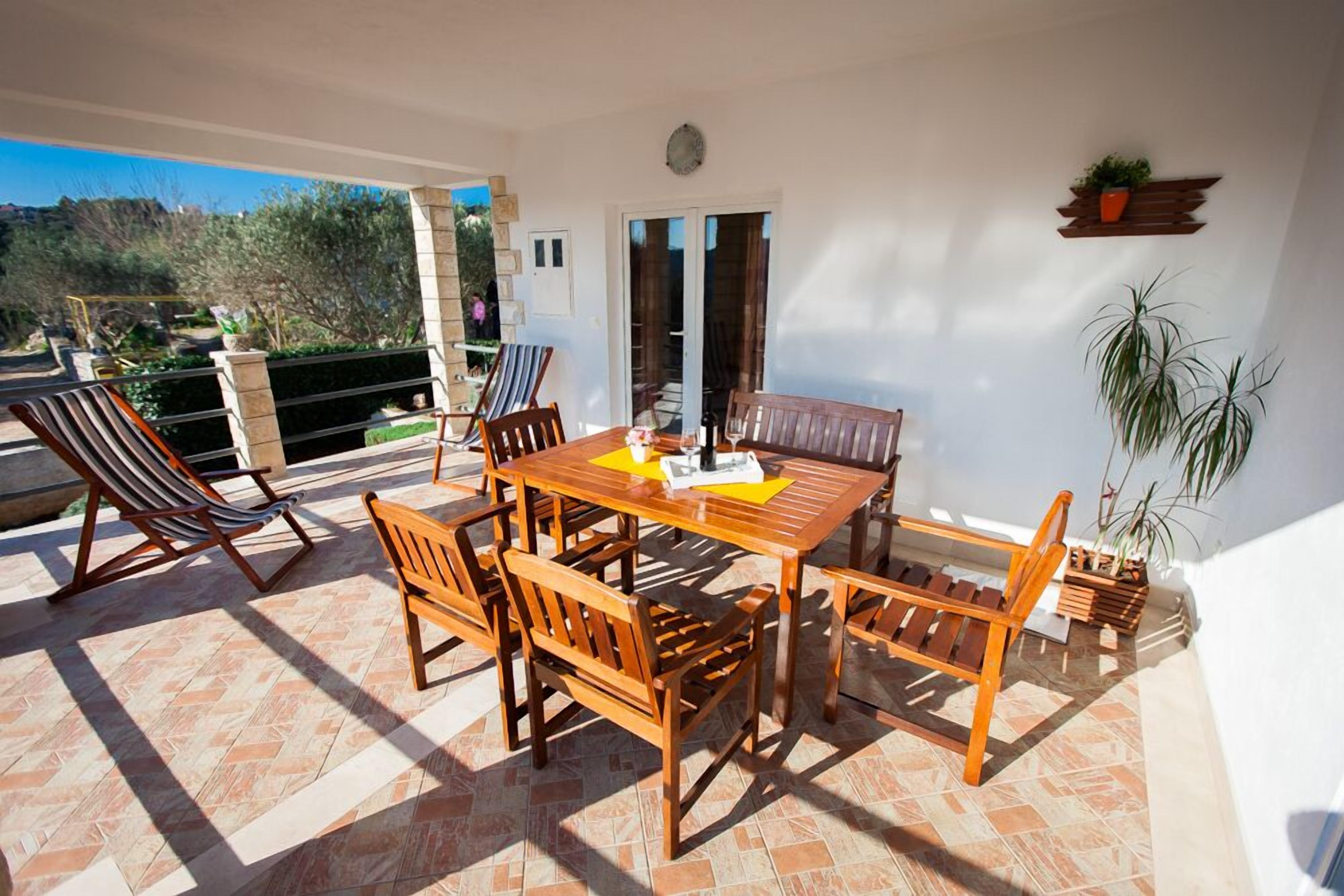 Um pátio ensolarado com mesa e cadeiras de madeira, uma poltrona estofada listrada e plantas em vasos.
