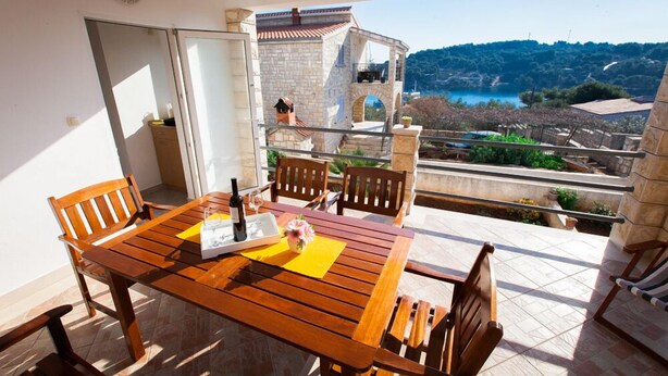 A wooden table with chairs on a balcony overlooking a scenic view of a stone building and a body of water. - Apartments Irene's Place (Solta)