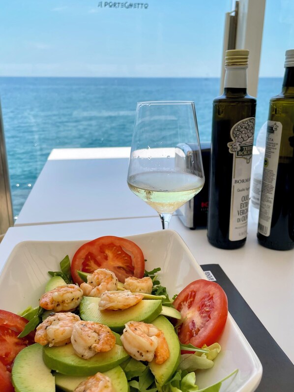 Een tafel met een witte schaal garnissalade, een glas witte wijn en twee flessen olijfolie.