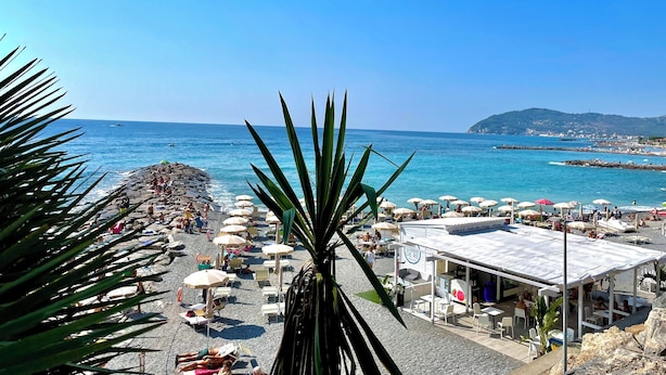 Una playa con sombrillas y tumbonas, un litoral rocoso y vistas al mar y colinas distantes.