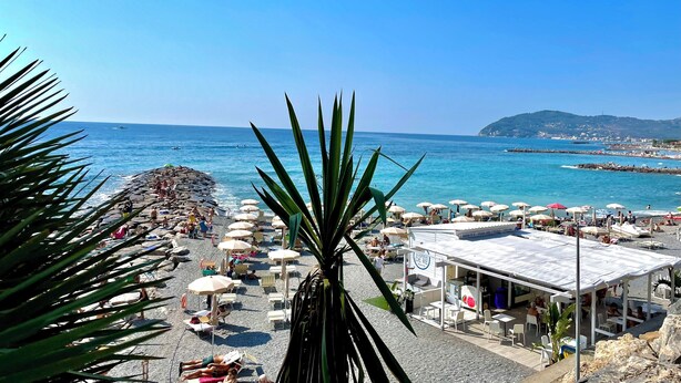 Une plage avec des parasols et des chaises longues, un rivage rocheux et une vue sur la mer et les collines lointaines.