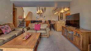 A spacious living room with a large sofa, a coffee table, and a television on a wooden cabinet. A staircase leads to another level, and there are wall decorations and a chandelier. - Spacious 3-Bedroom + Loft Condo at Timber Run – Perfect for Families and Groups! (Steamboat Springs)