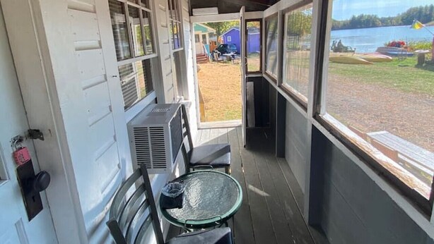 A covered porch with a glass table and chairs, a view of a lake, and a building in the background. - Lakefront  Yellow Cottage (Northwood)