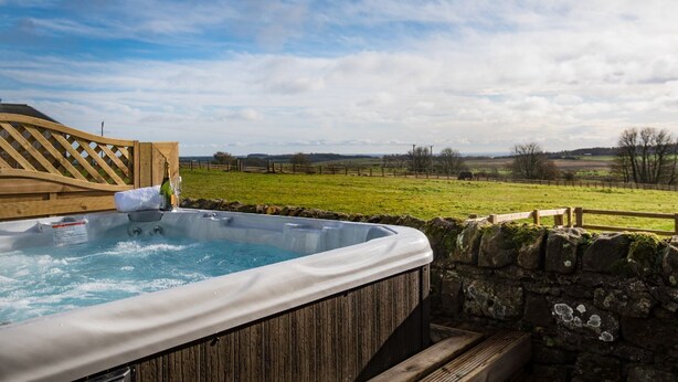 Een hottub met een houten terras en een stenen muur, gelegen in een landelijk gebied met velden en bomen.