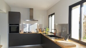 A modern kitchen with dark cabinetry, a stainless steel range hood, a built-in oven, and a large window providing natural light. - Beautiful house with heated pool overlooking Barjac (Barjac)