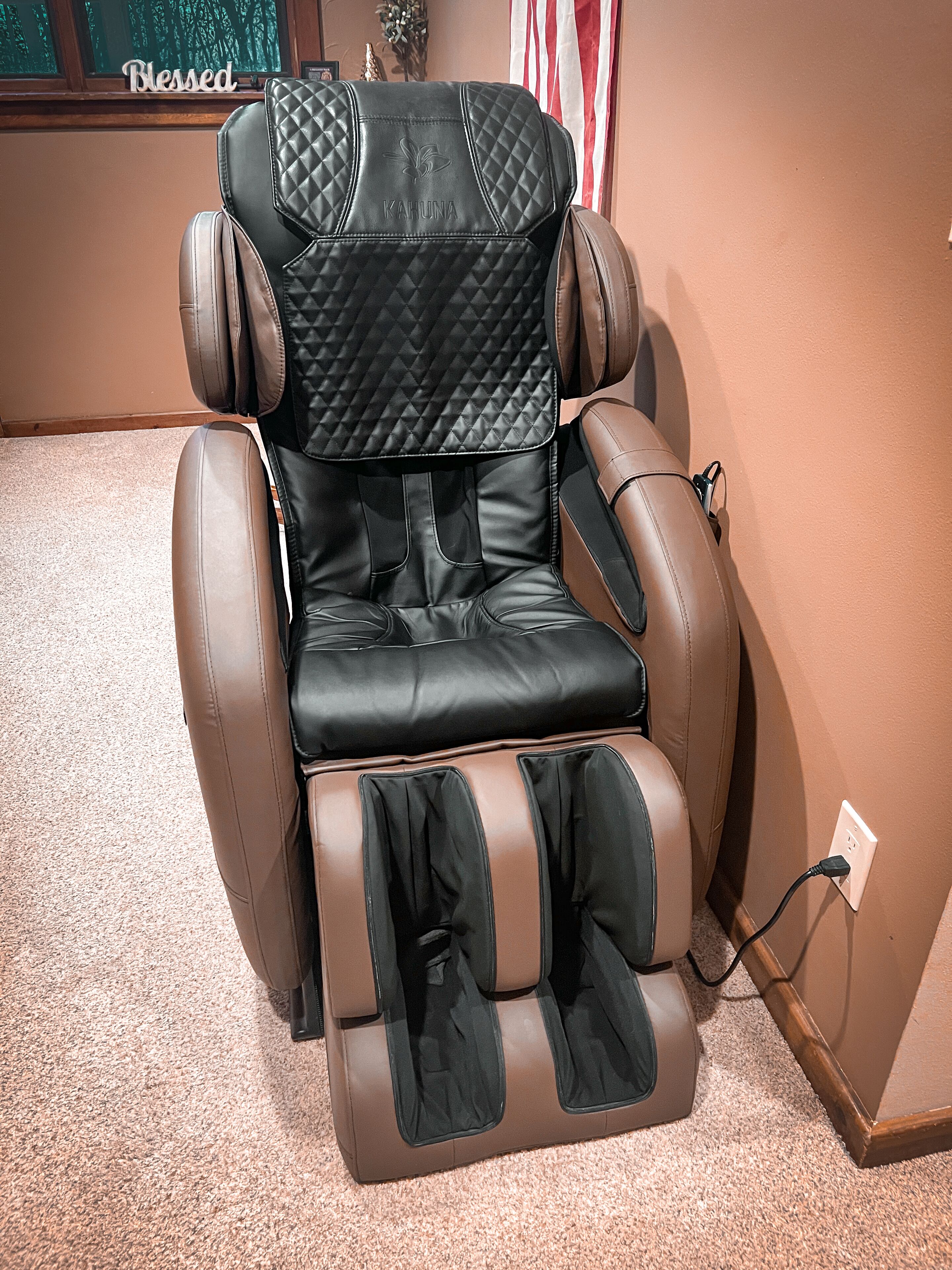 A massage chair with a quilted backrest and footrest, positioned in a room with a carpeted floor and a wall-mounted power outlet.