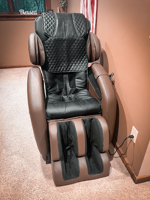 A massage chair with a quilted backrest and footrest, positioned in a room with a carpeted floor and a wall-mounted power outlet. - Rustic Charm: Pewaukee Lake Cabin, Sauna & Hot Tub (Pewaukee)