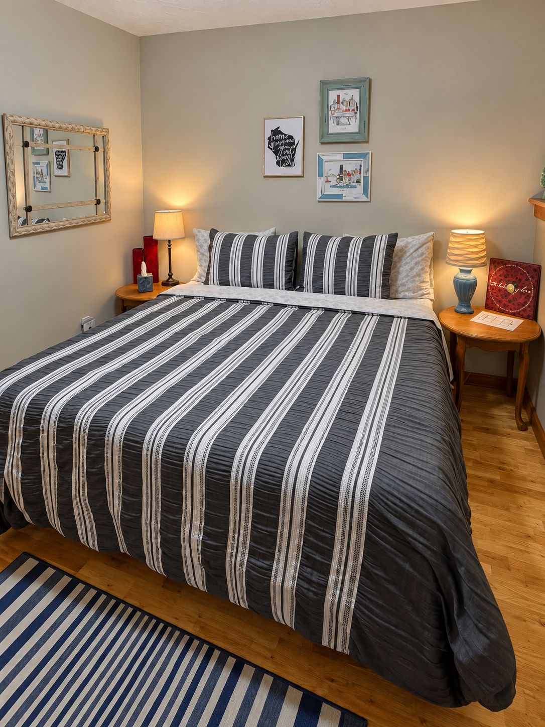 Een opgemaakt bed met gestreepte lakens, een nachtkastje met een lamp, en kunstwerken in lijsten aan de muur.