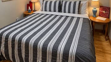 Een opgemaakt bed met gestreepte lakens, een nachtkastje met een lamp, en kunstwerken in lijsten aan de muur.