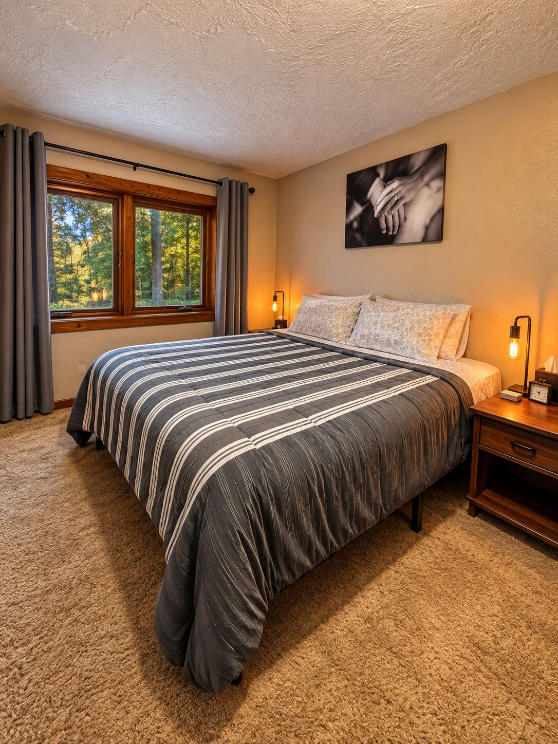 A bedroom with a large bed, a window with curtains, a bedside table with a lamp, and a wall-mounted picture.