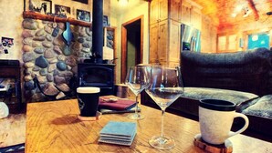 Ein gemütliches Zimmer mit Steinwand, Holzmöbeln, Kamin und Tisch mit Kaffeetasse, Weingläsern und Notizbuch.