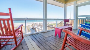 A wooden deck with a view of the beach, a red rocking chair, a pink table, and a blue bench.