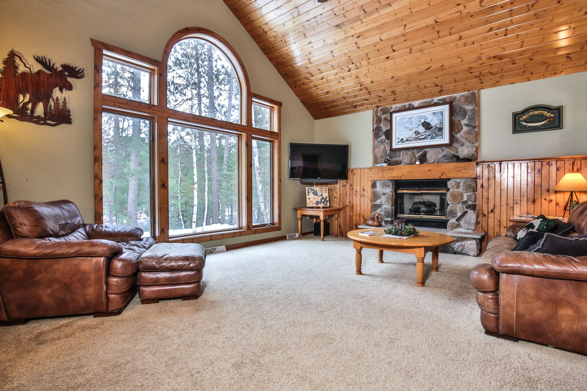 A cozy living room with leather sofas, a stone fireplace, large windows, and a television.