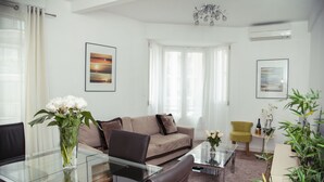 Una sala de estar moderna con una mesa de comedor de vidrio, un sofá color crema y un sillón amarillo. Hay dos ventanas amplias con cortinas translúcidas, un candelabro y un cuadro enmarcado en la pared.