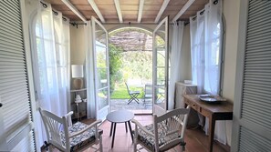 A room with a view of a garden, a table with chairs, a lamp, and a small table with a bowl.