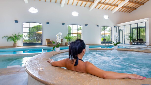 En kvinde slapper af i en jacuzzi med en pool og store vinduer i baggrunden.