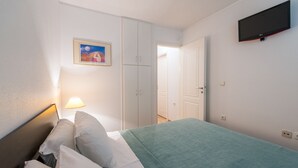 Une chambre à coucher comprenant un lit, une table de chevet, une lampe, une armoire, un téléviseur et une porte donnant sur une autre pièce.