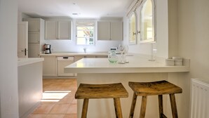 A modern kitchen with white cabinetry, a large island with stools, and a window with patterned curtains. - 4 BEDROOM VILLA - PRIVATE POOL AND GARDEN (N°348) (La Motte)