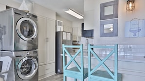 A compact kitchen with a washing machine, a refrigerator, and a teal wooden chair. - 2nd Floor @ THE SUMMIT 1BR 1.5BA W/ BEACH CHAIRS INCLD. (Panama City Beach)