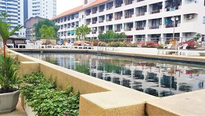 A swimming pool with lounge chairs and potted plants, surrounded by a building with balconies and air conditioning units. - Plaza Residence Jomtien Beach - Large Studio Condo (Pattaya)
