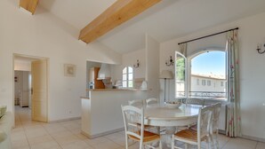 Un comedor con una mesa redonda y sillas blancas, una cocina al fondo y vistas de edificios a través de una ventana.