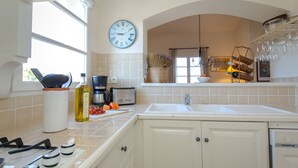 A kitchen with a gas stove, a coffee maker, and a sink. There is a clock on the wall and a basket of vegetables on the counter. - 2-BEDROOM APARTMENT WITH TERRACE (N°555) (La Motte)