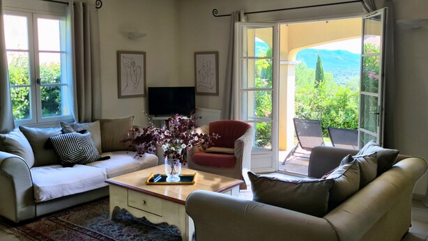 A living room with a sofa, armchair, and a coffee table with a vase of flowers. There is a television on a stand and a view of greenery outside. - 3 BEDROOM TOWNHOUSE WITH GARDEN (N°528) (La Motte)