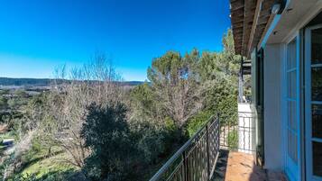 Un balcon donnant sur un paysage vallonné, une pergola en bois et un bâtiment avec des portes bleues.