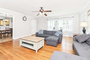 A living room with a ceiling fan, a grey sofa, a wooden coffee table, and a dining area with chairs and a table. - Charming 3BR Home in Rossville, GA Family Retreat (Rossville)