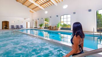 Une piscine intérieure avec vue sur l’extérieur, un plafond avec des poutres en bois et un espace détente avec des fauteuils et des plantes.