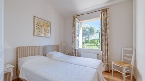 Ein Schlafzimmer mit zwei Betten, einem Sessel, einem Fenster mit Vorhängen und einem gerahmten Bild an der Wand.