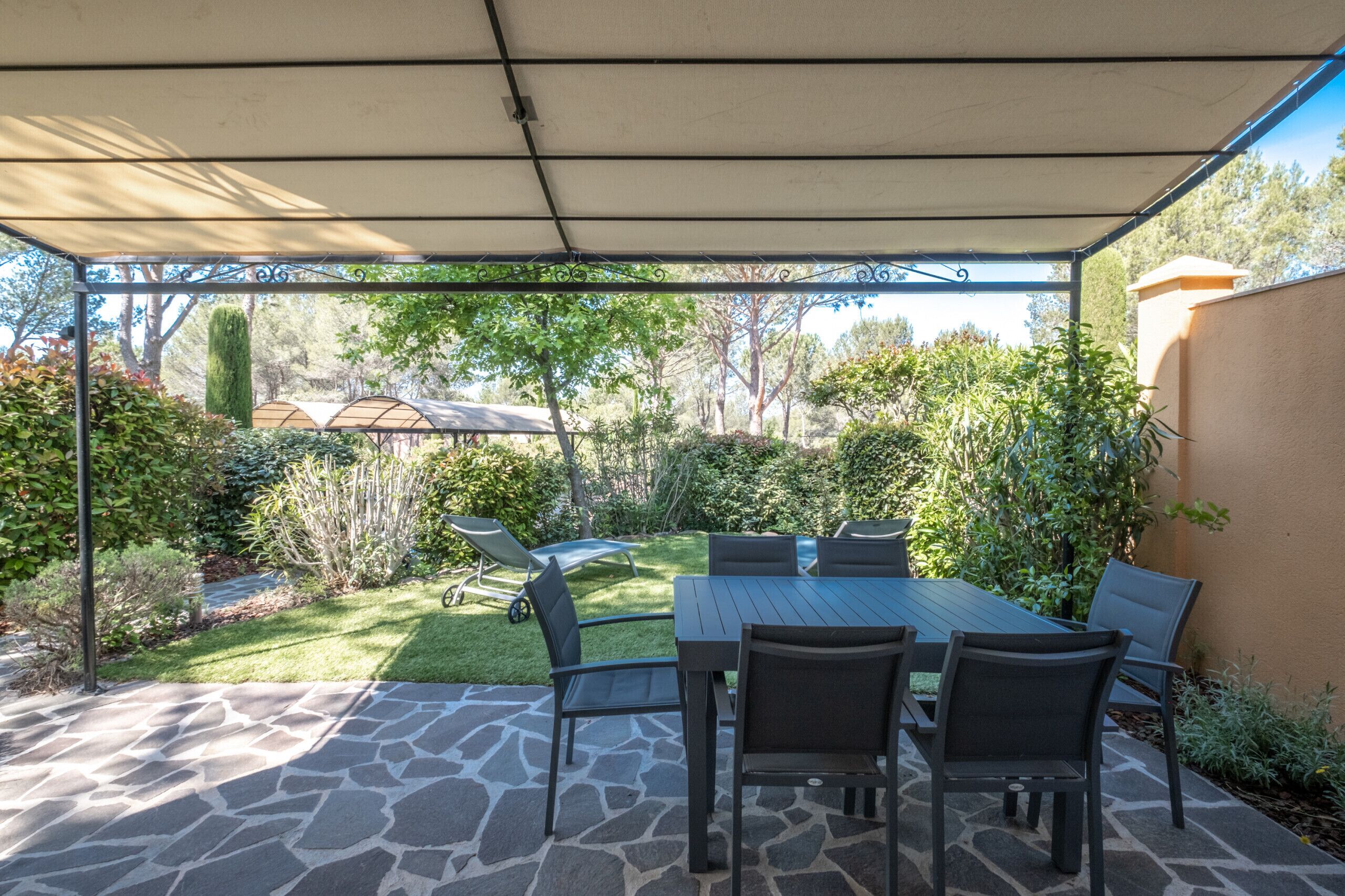 An outdoor patio area with a stone floor, a covered table with chairs, and a view of a garden with various plants and trees.