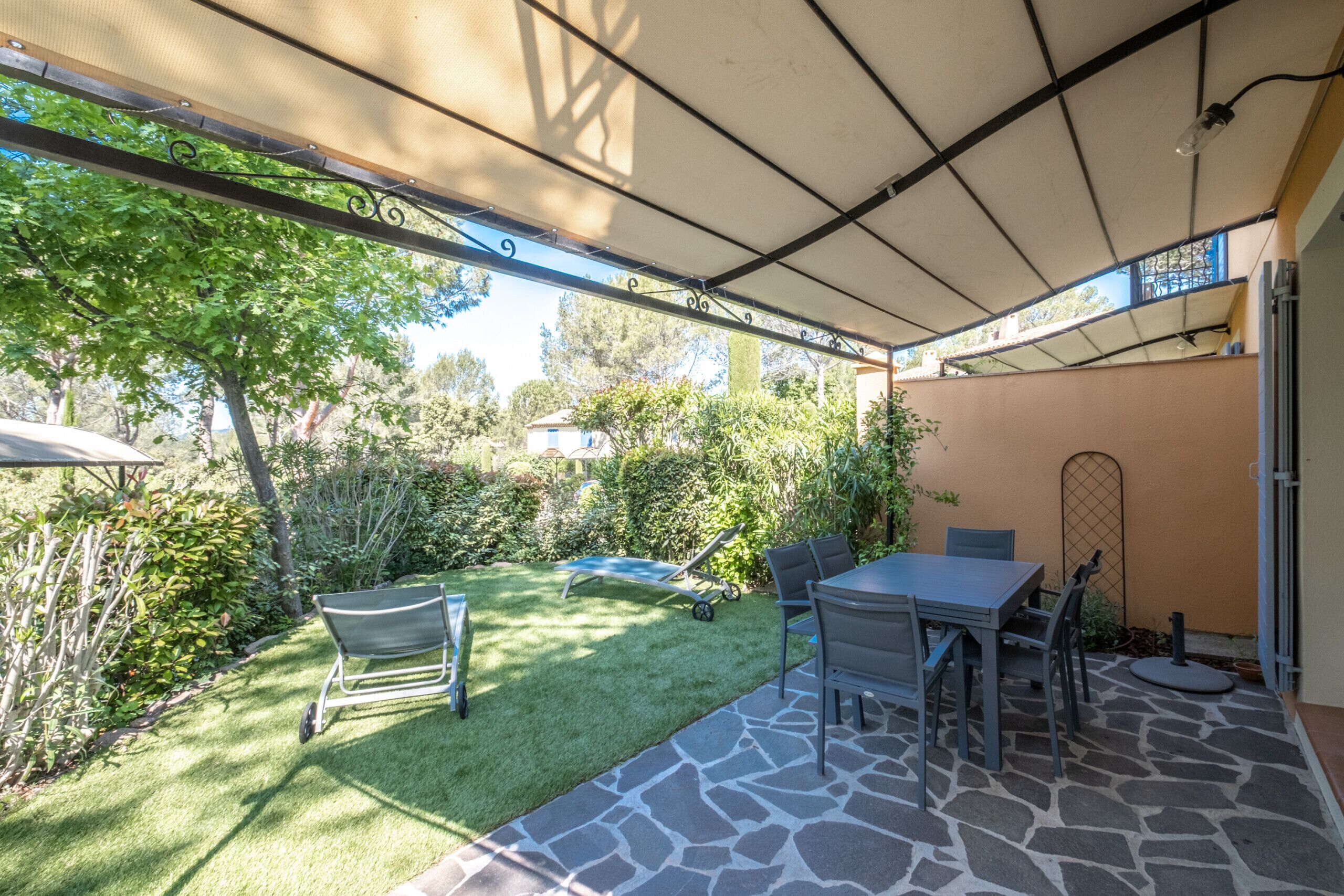 Un espace extérieur aménagé en terrasse, avec un sol en pierre, une table et des chaises, une pelouse et un coin salon abrité.