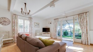 Ein Wohnzimmer mit Sofa, Fernseher, Esstisch und einem großen Fenster mit Vorhängen.