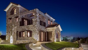 A large stone house with a stone facade, multiple windows, and a well-lit entrance. - Palazzo Villa, World 5th Awarded Villa (Zakynthos)