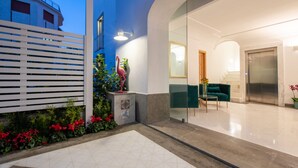 Una hall d'hotel moderna con pavimento in piastrelle geometriche, una reception bianca, un divano verde e una scala che conduce a un ascensore.