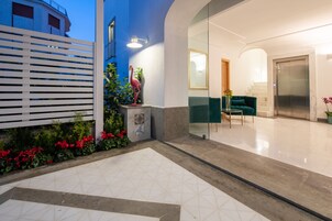 Un vestíbulo de hotel moderno con piso de baldosas geométricas, un mostrador de recepción blanco, un sofá verde y una escalera que conduce a un ascensor.