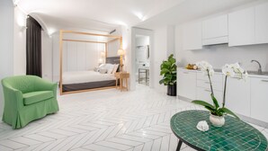 Una moderna camera d'albergo con un letto, una poltrona verde, un tavolo da pranzo con un'orchidea in vaso e un angolo cottura con pensili bianchi e un lavandino.