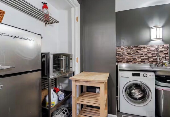 A compact kitchen with a stainless steel refrigerator, a microwave, a wooden stool, and a washing machine.