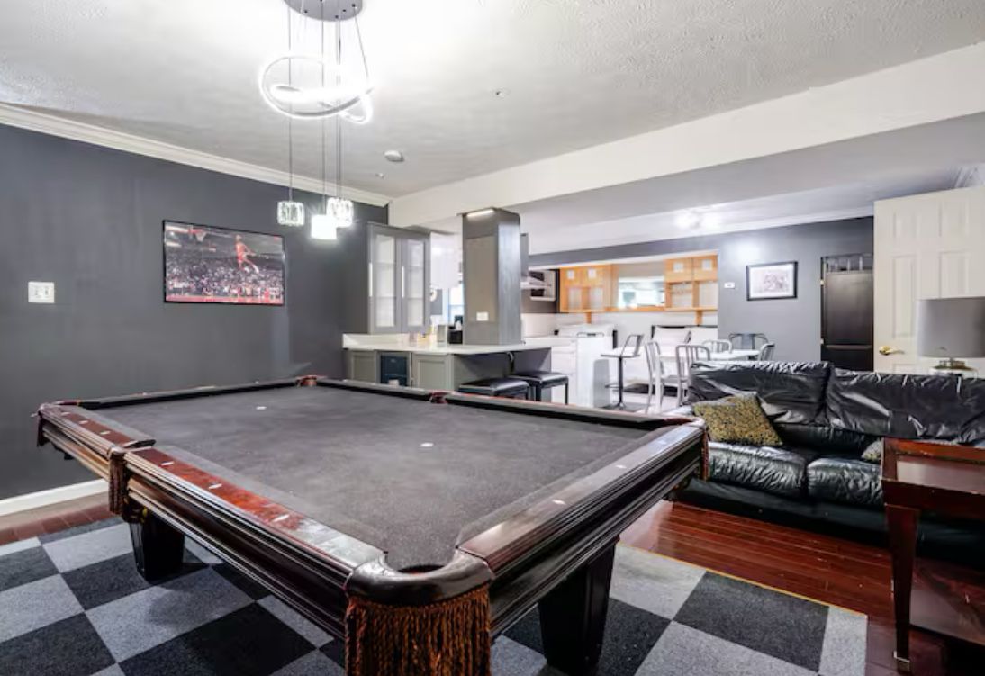 A pool table in a room with a checkered floor, a leather couch, and a kitchen area in the background.