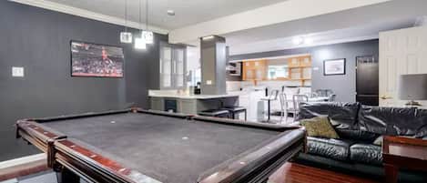 Una mesa de billar en una habitación con suelo a cuadros, un sofá de cuero y una zona de cocina al fondo.