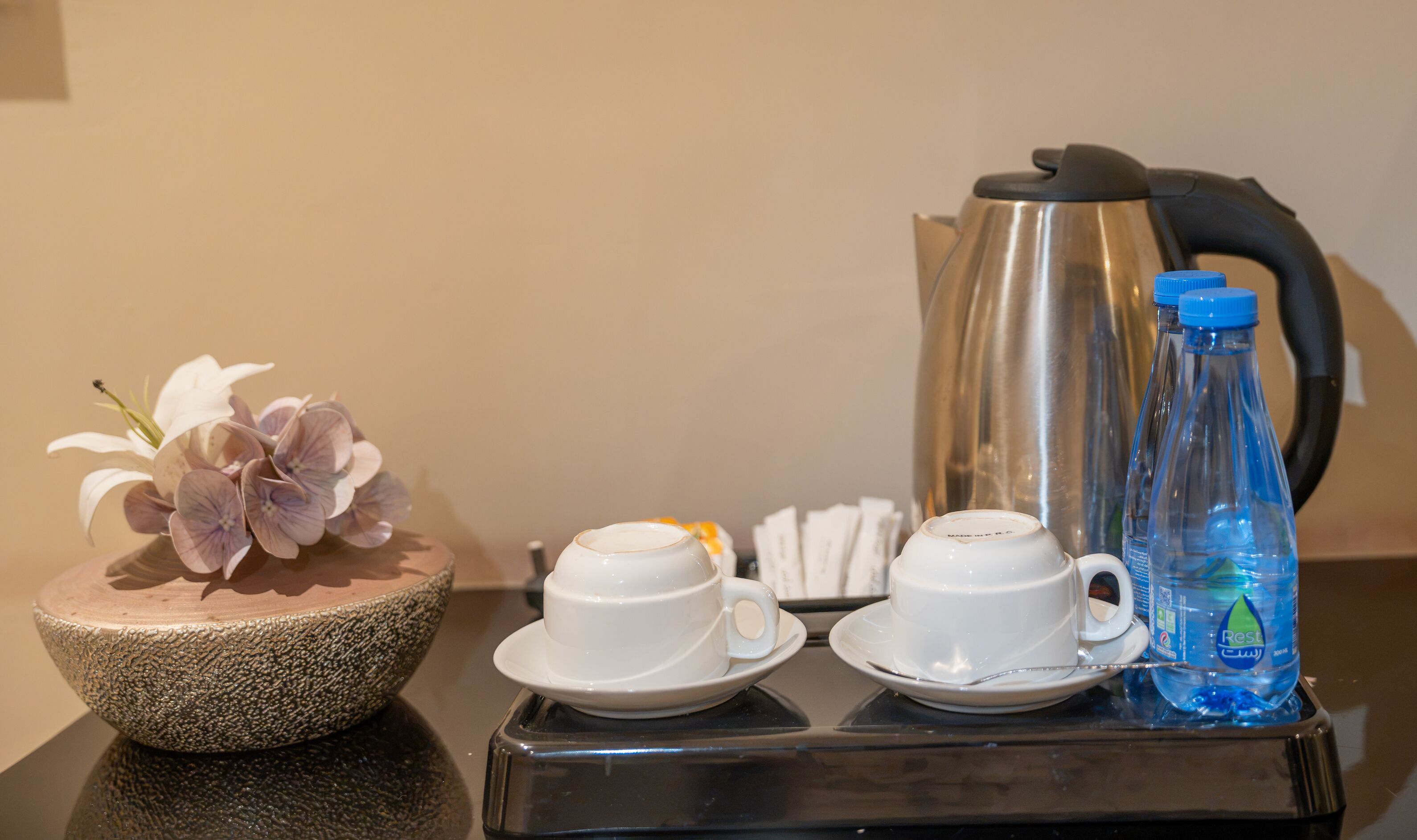 A table with two white cups, a water bottle, a kettle, and a decorative bowl with flowers.