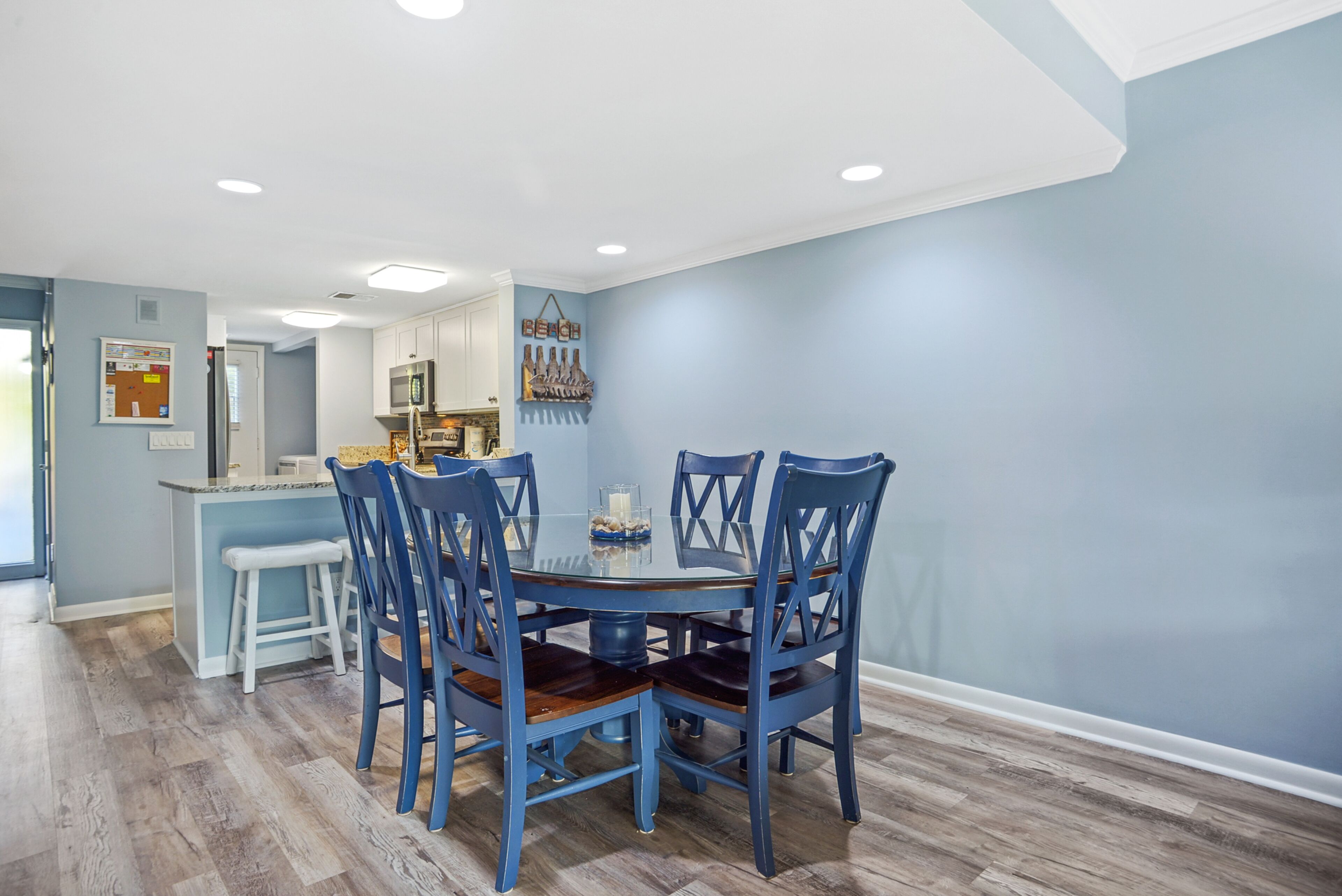 A dining area with a round table and blue chairs, a kitchen in the background, and a light blue wall.