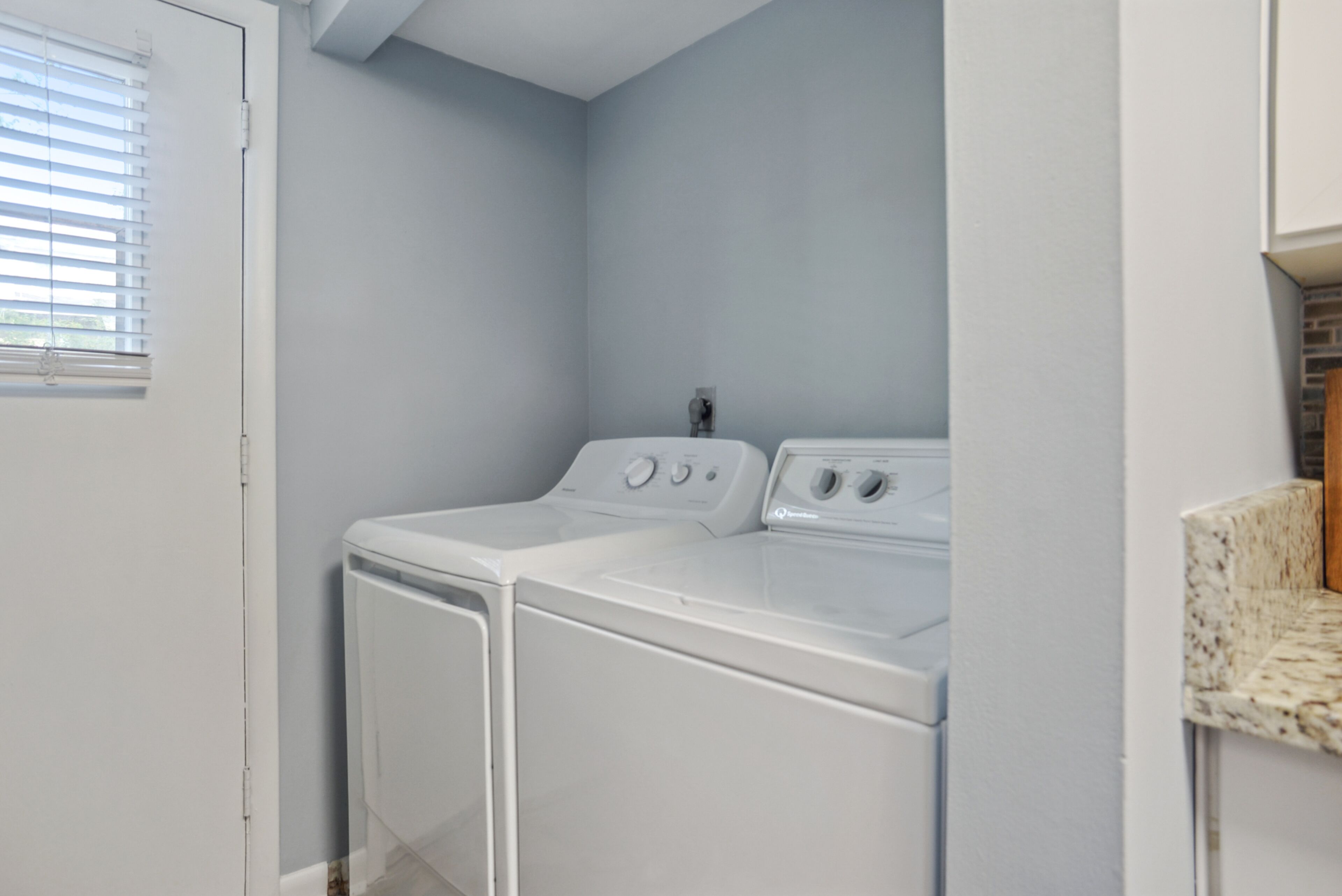 Une buanderie équipée d’un lave-linge et d’un sèche-linge à chargement frontal, d’une petite fenêtre avec des stores et d’un plan de travail avec un évier.