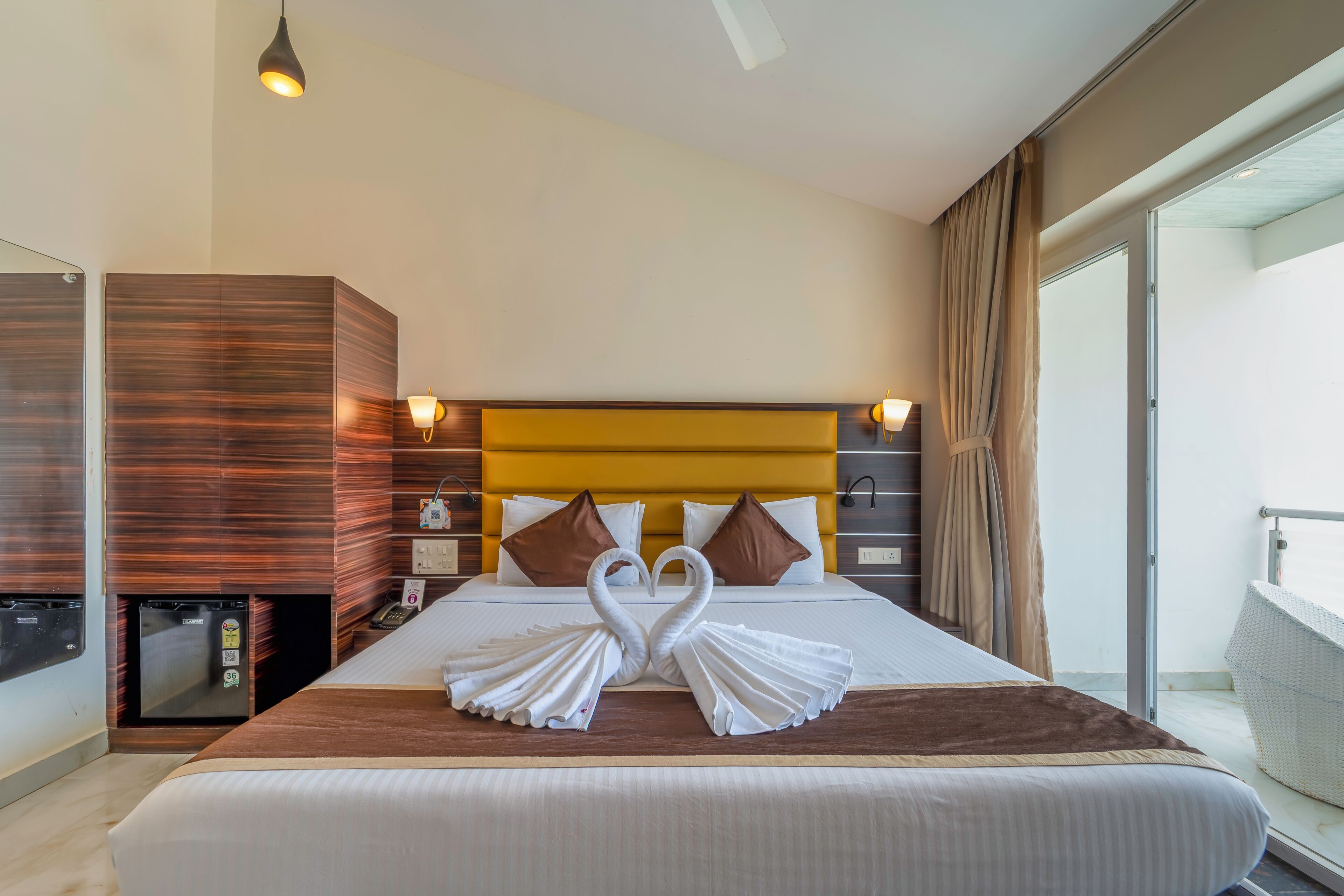 A hotel room with a bed, a minibar, a desk, and a balcony with curtains.