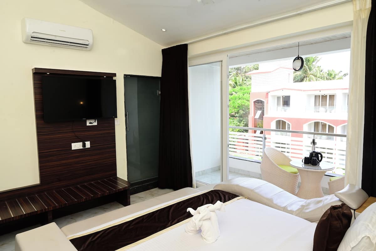 A hotel room with a bed, a TV mounted on a wooden panel, a window with a view of buildings and greenery, and a small table with a coffee maker.