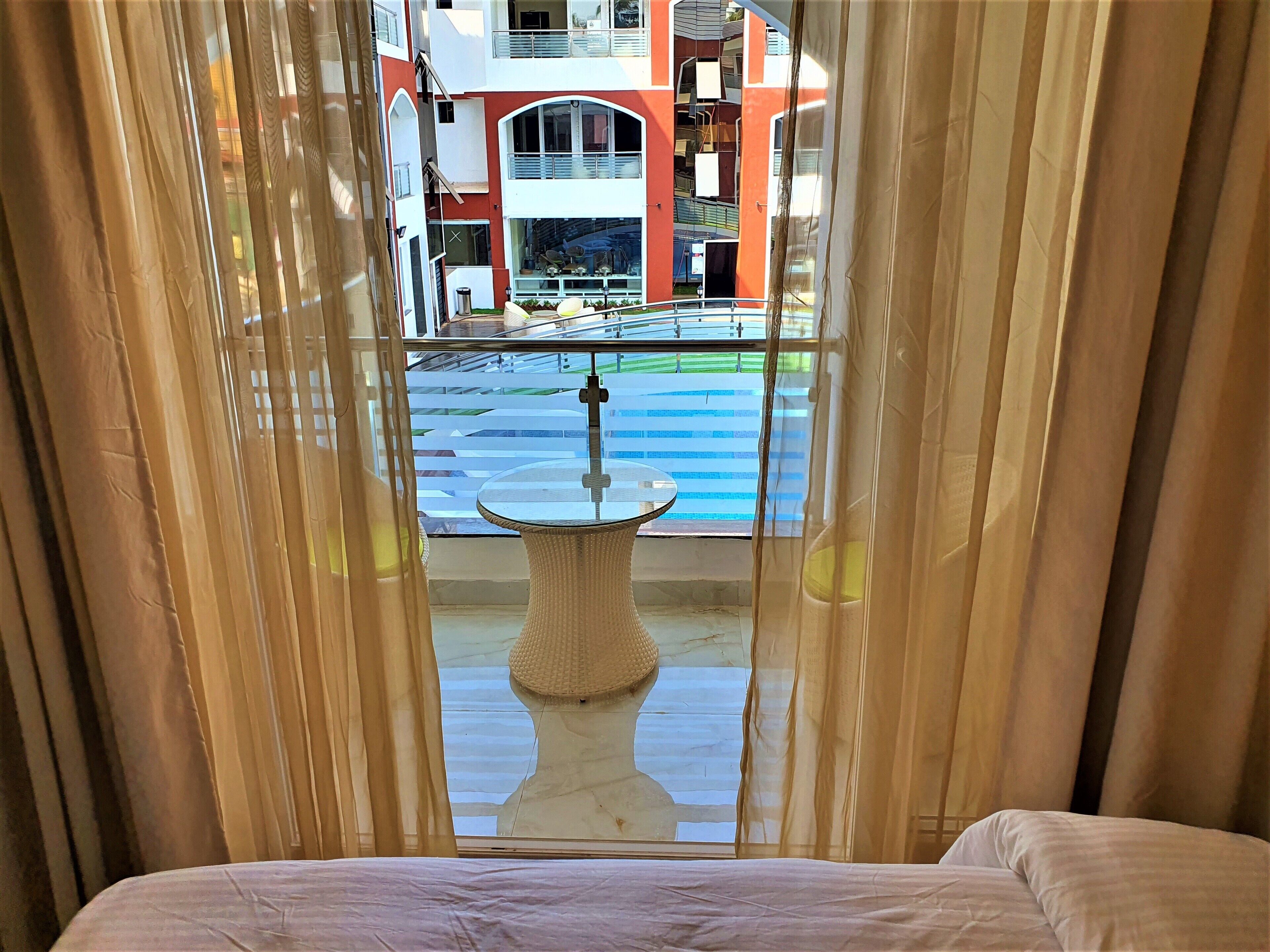 A hotel room with a bed, a window with sheer curtains, and a view of a pool and buildings outside.
