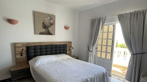 A bedroom with a wooden headboard, a bed with a white bedspread, a painting on the wall, and a view of the outdoors through a window with curtains.
